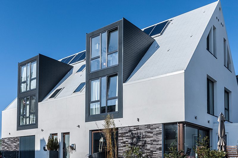 Weißes Wohnhaus mit steilem Satteldach und dunklen Gauben vor blauem Himmel